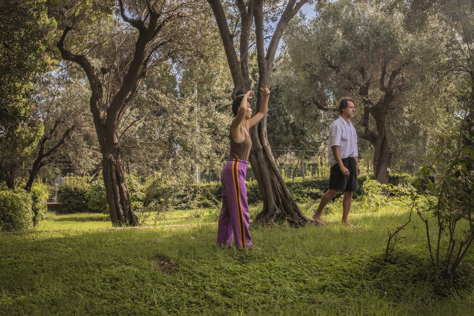 Two dancers among olive trees, one with arms raised, the other walking barefoot