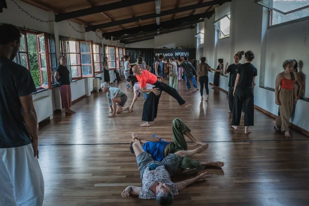 A long bright studio with open windows, dancers moving and resting on the wooden floor